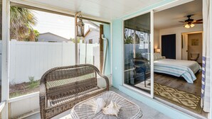 A covered patio with a wicker chair and a round table, a bedroom with a bed and a ceiling fan, and a view of a white fence and palm trees. - Breezy Englewood Condo w/ Community Pool! (Englewood)