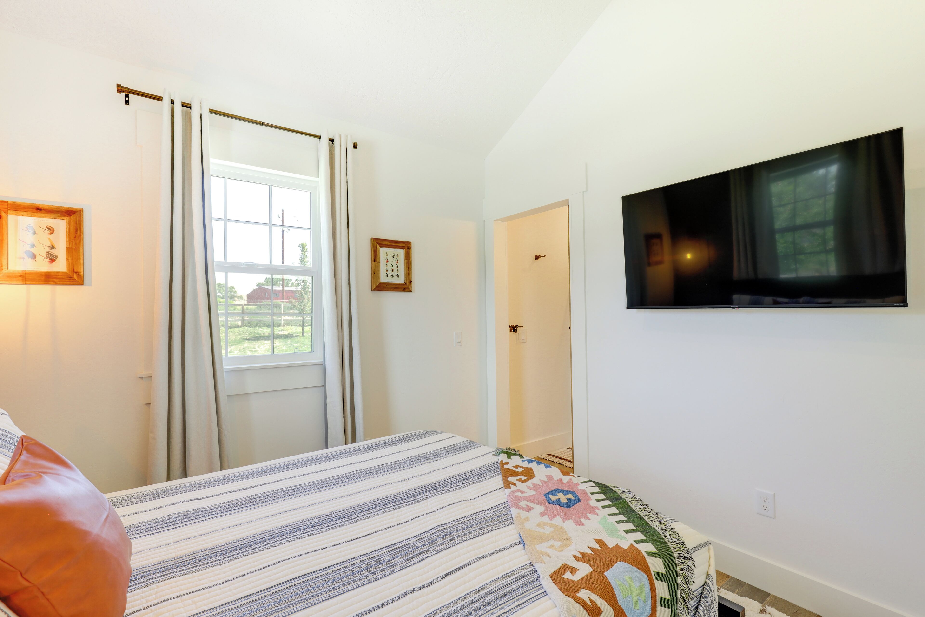Un dormitorio con una cama, un televisor montado en la pared, una ventana con cortinas y una puerta.