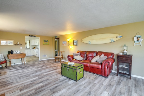 A living room with a red leather couch, a green trunk, and a surfboard on the wall. - Winchester Bay Vacation Rental Near Dunes & Atv! (Reedsport)