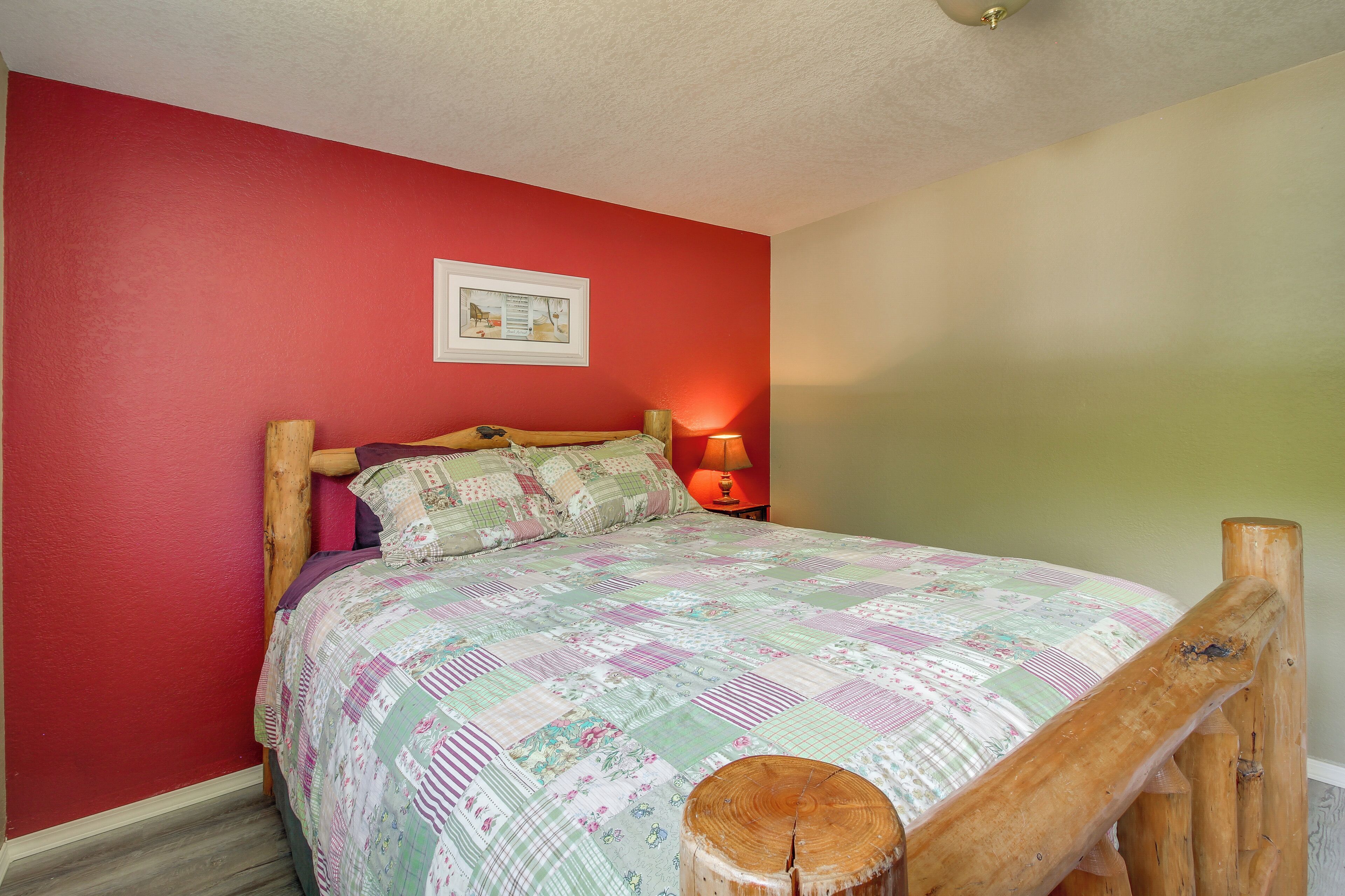 A bedroom with a red wall, a wooden bed frame, a quilted bedspread, a nightstand with a lamp, and a framed picture on the wall.