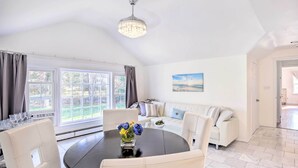Ein geräumiges Wohnzimmer mit einem runden schwarzen Tisch, weißen Ledersesseln, einem weißen Sofa und einem großen Fenster mit Vorhängen.