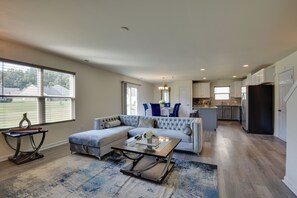 A spacious living room with a sectional sofa, a glass coffee table, and a view of the backyard through large windows. - Spacious Gastonia Home w/ Pool Access! (Gastonia)
