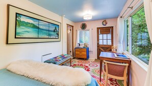 Un dormitorio con cama, escritorio, silla, cómoda, ventana con cortinas y un cuadro en la pared.
