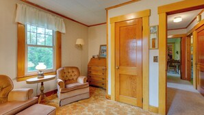 A room with a large wooden door, a window with a white valance, a tufted armchair, a small table with a lamp, and a dresser. - Cape House in Farmington w/ Shared Outdoor Pool! (Farmington)