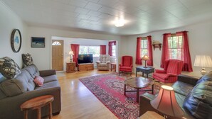 A spacious living room with a large sofa, a coffee table, and two armchairs. There is a television on a wooden stand, and a red rug on the floor.