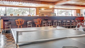 A bar area with wooden stools, a ping pong table, and a view of the outdoors through large windows.