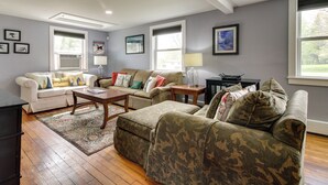 A living room with a white sofa, a beige sofa, and a camouflaged sofa. There are two windows, a lamp, and a rug. - Vermont Vacation Rental: Hot Tub, Near Ski Resorts (Mount Holly)