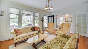 A spacious living room with a beige sofa, a floral sofa, and a matching armchair. There are large windows with blinds, a chandelier, and a door with glass panels. - Elizabeth City Vacation Rental: Gazebo w/ Hot Tub! (Elizabeth City)