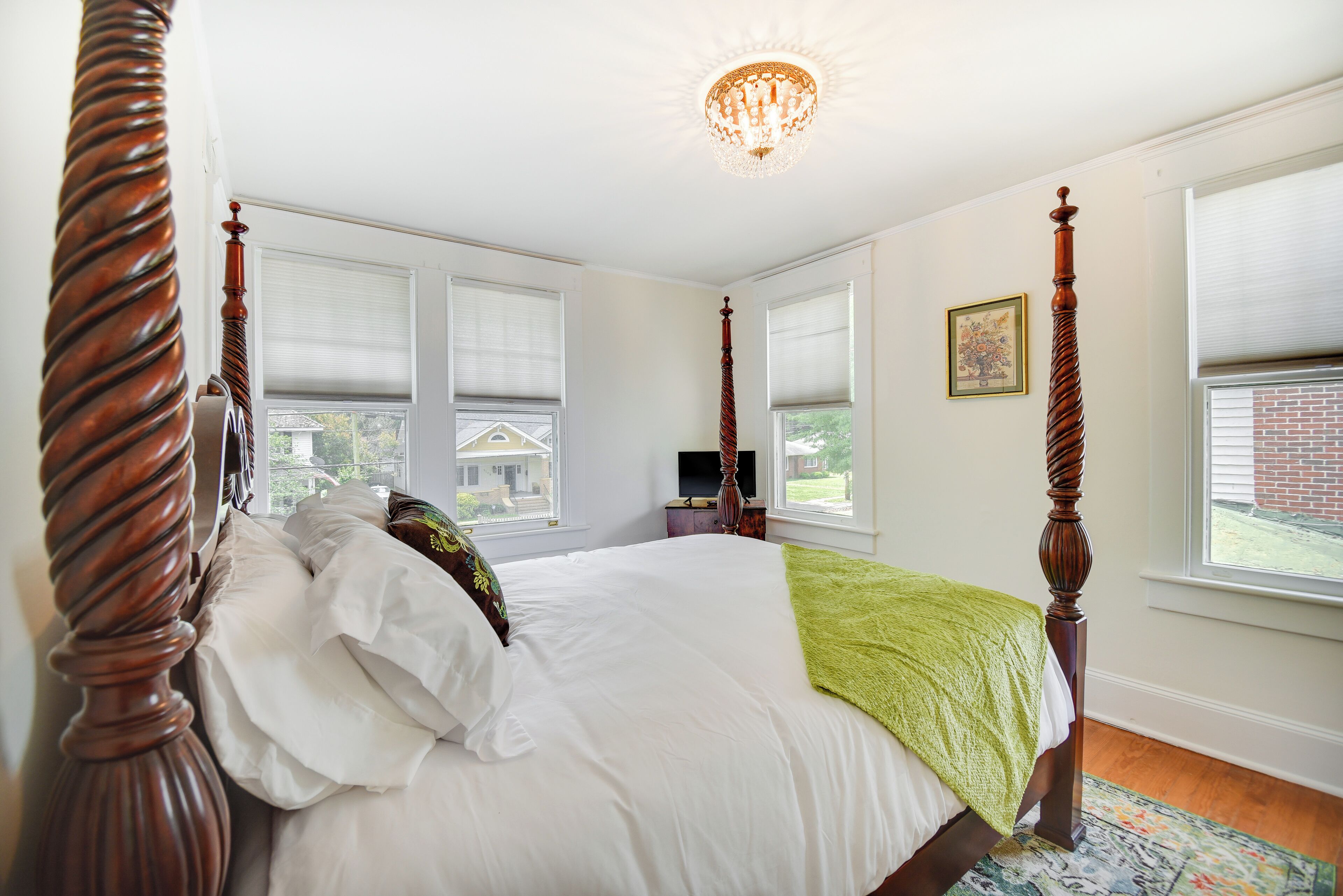 A four-poster bed with white linens and a green throw blanket, a chandelier, and a view of a house through the windows.