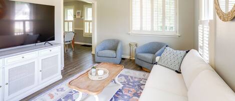 Un salon doté d’un ventilateur de plafond, d’un téléviseur à écran plat, d’un canapé blanc, de deux fauteuils, d’une table basse et d’un tapis à motifs.