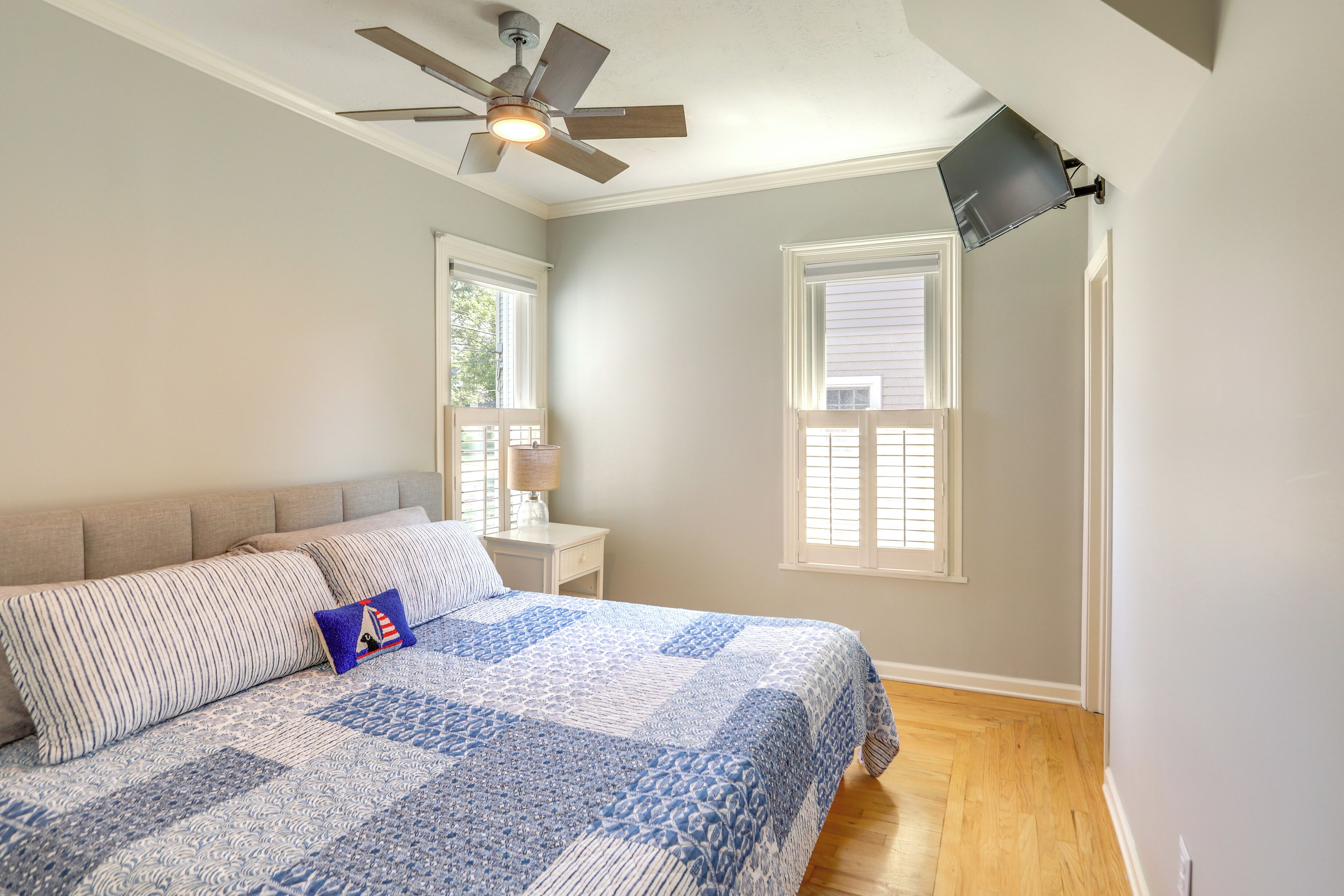 A bedroom with a bed, a ceiling fan, a nightstand, a lamp, and two windows.
