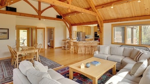 A spacious living area with a high wooden ceiling, a dining table with chairs, a sofa, and a kitchen in the background.