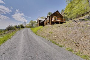 Une cabane en bois dotée d’un balcon, située sur un chemin de gravier, entourée d’arbres et bordée d’une colline couverte d’herbe.