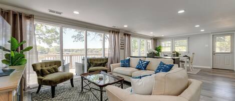 A spacious living room with a beige sofa, a green armchair, and a coffee table. There is a large window providing a view of the outdoors.