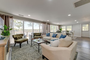 A spacious living room with a beige sofa, a green armchair, and a coffee table. There is a large window providing a view of the outdoors.