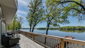 Eine Holzterrasse mit Gartenmöbeln, mit Blick auf einen stillen See, Bäume und bewaldete Hügel in der Ferne.