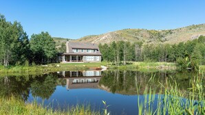 A house with a large porch, a pond in front, and a hilly landscape in the background. - Tranquil Wyoming Cabin Near Uintas - 105 Acres! (Evanston)