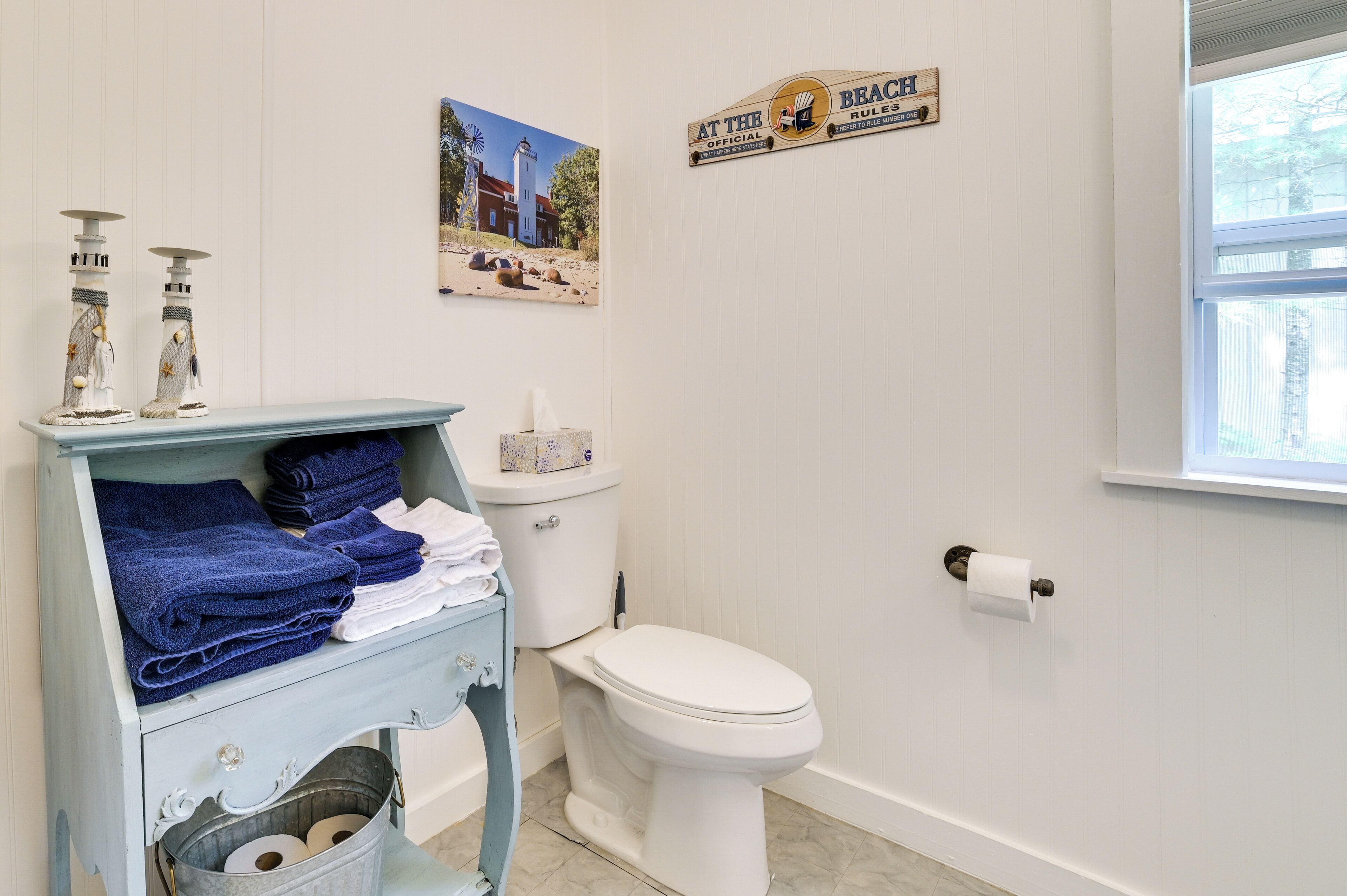 A bathroom with a white toilet, a blue vanity with towels, a bucket, and a sign that reads "At the Official Brac'h Rules."