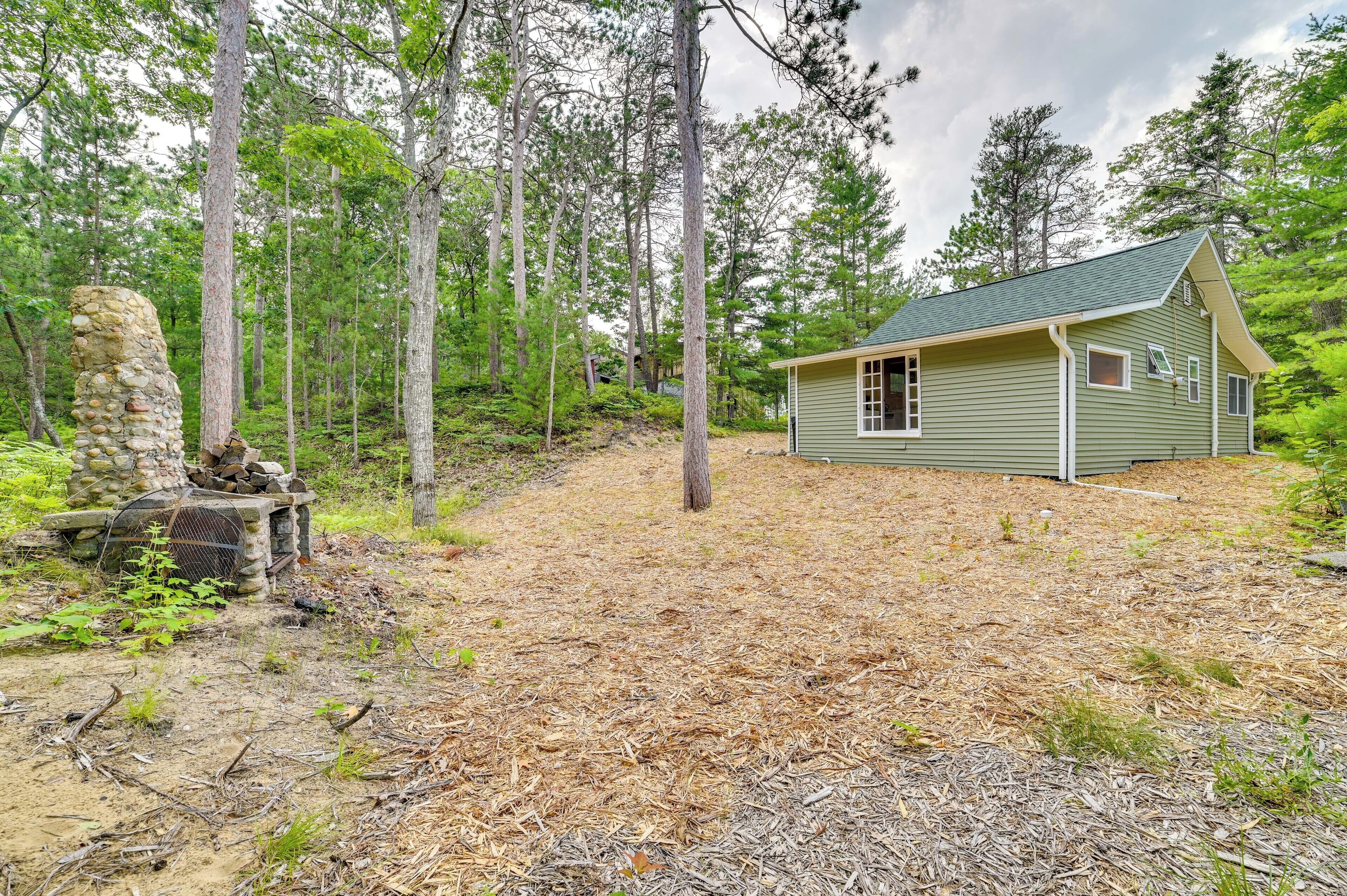 Une petite cabane avec un toit vert et des bordures blanches, entourée d’une zone boisée avec de grands arbres et une structure en pierre sur la gauche.
