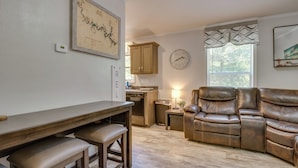 A living room with a brown leather sofa, a dining table with benches, a kitchen area, and a wall clock. - Oakland Oasis: Serene Retreat in the Ozarks (Oakland)
