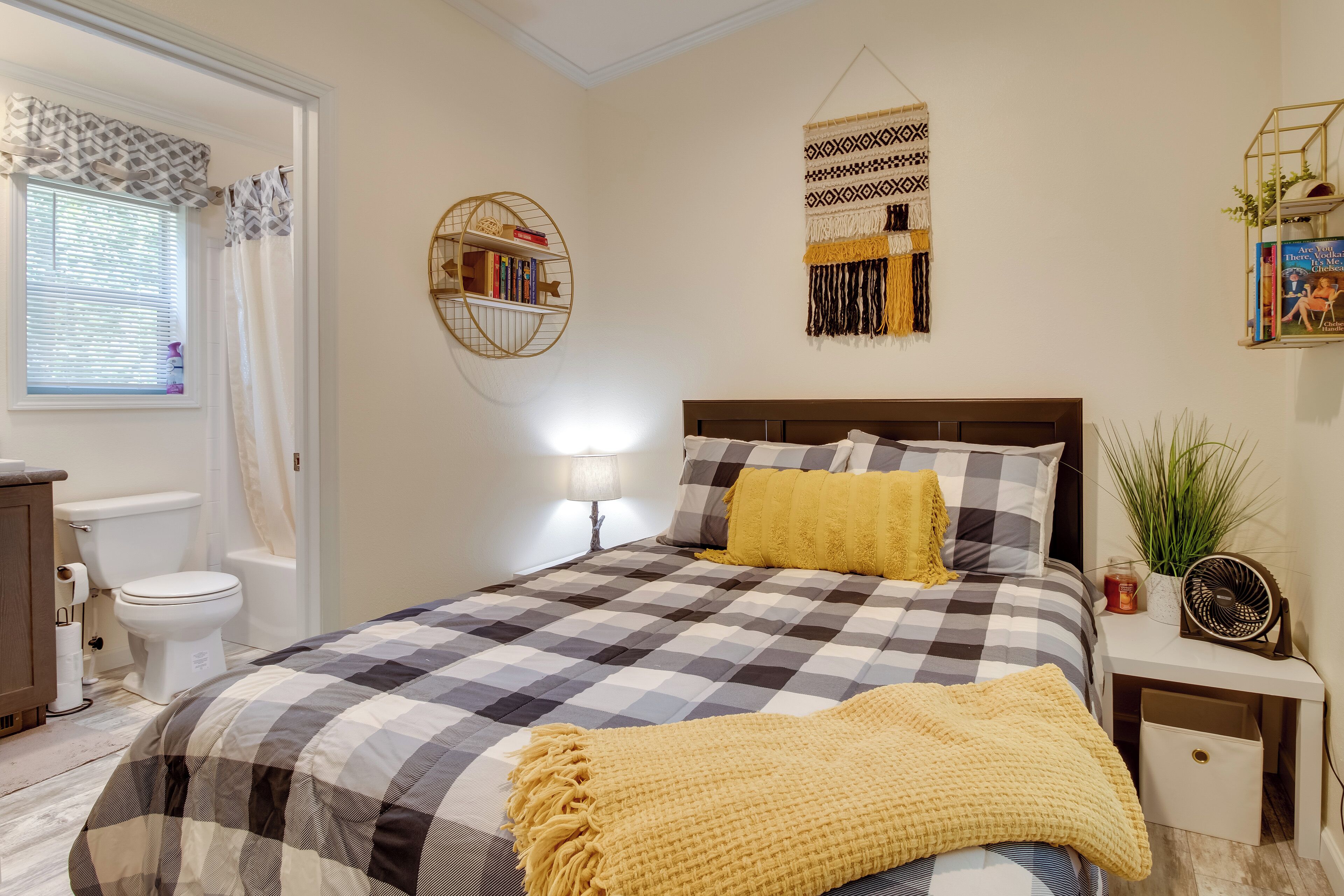 Ein Schlafzimmer mit karierter Bettdecke, einem Nachttisch mit Lampe, einem an der Wand montierten Regal mit Büchern und einem dekorativen Wandbild.