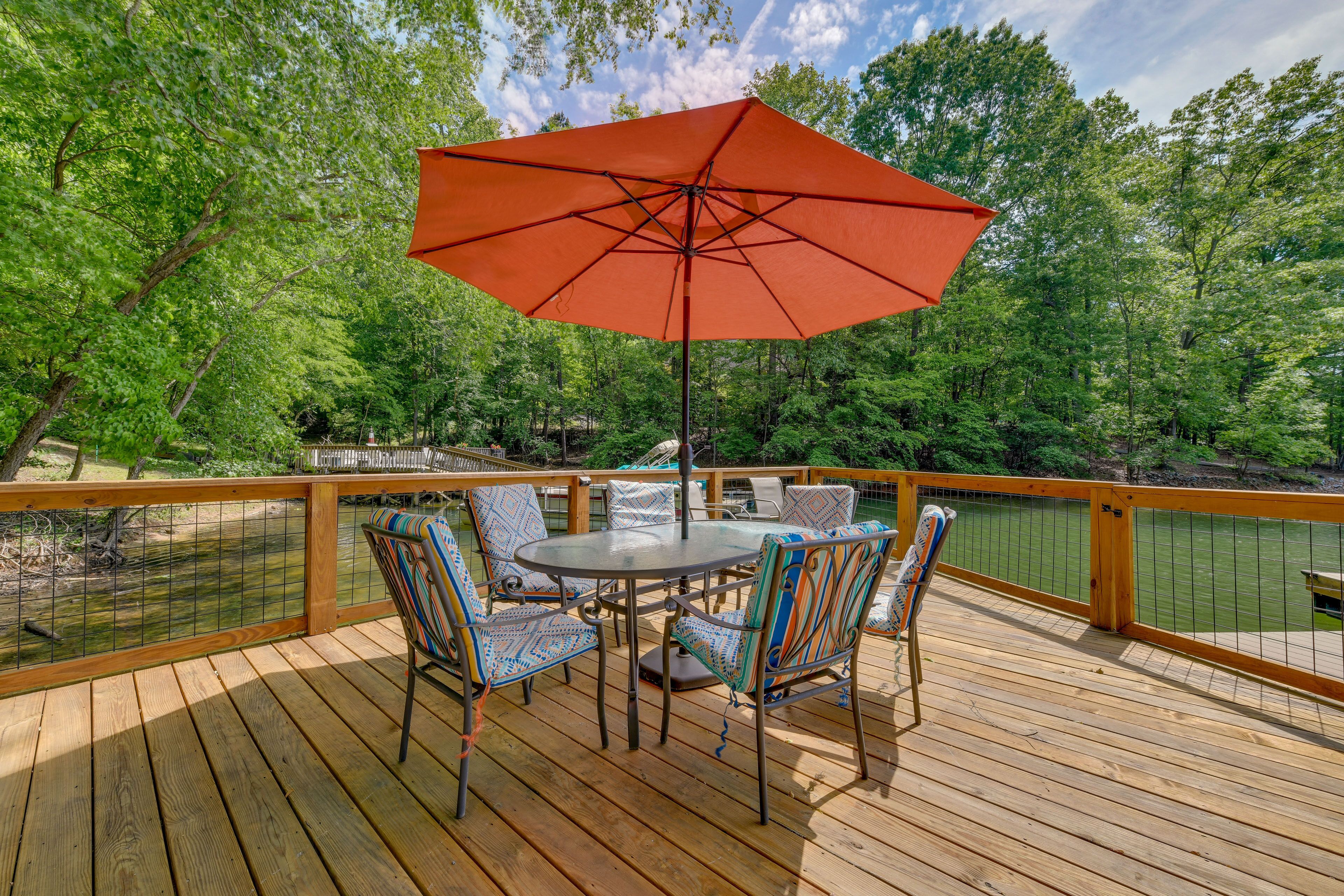 A wooden deck with a round table and chairs, an orange umbrella, and a view of a lake surrounded by trees.