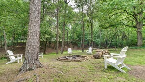 A forested area with a fire pit and white Adirondack chairs.