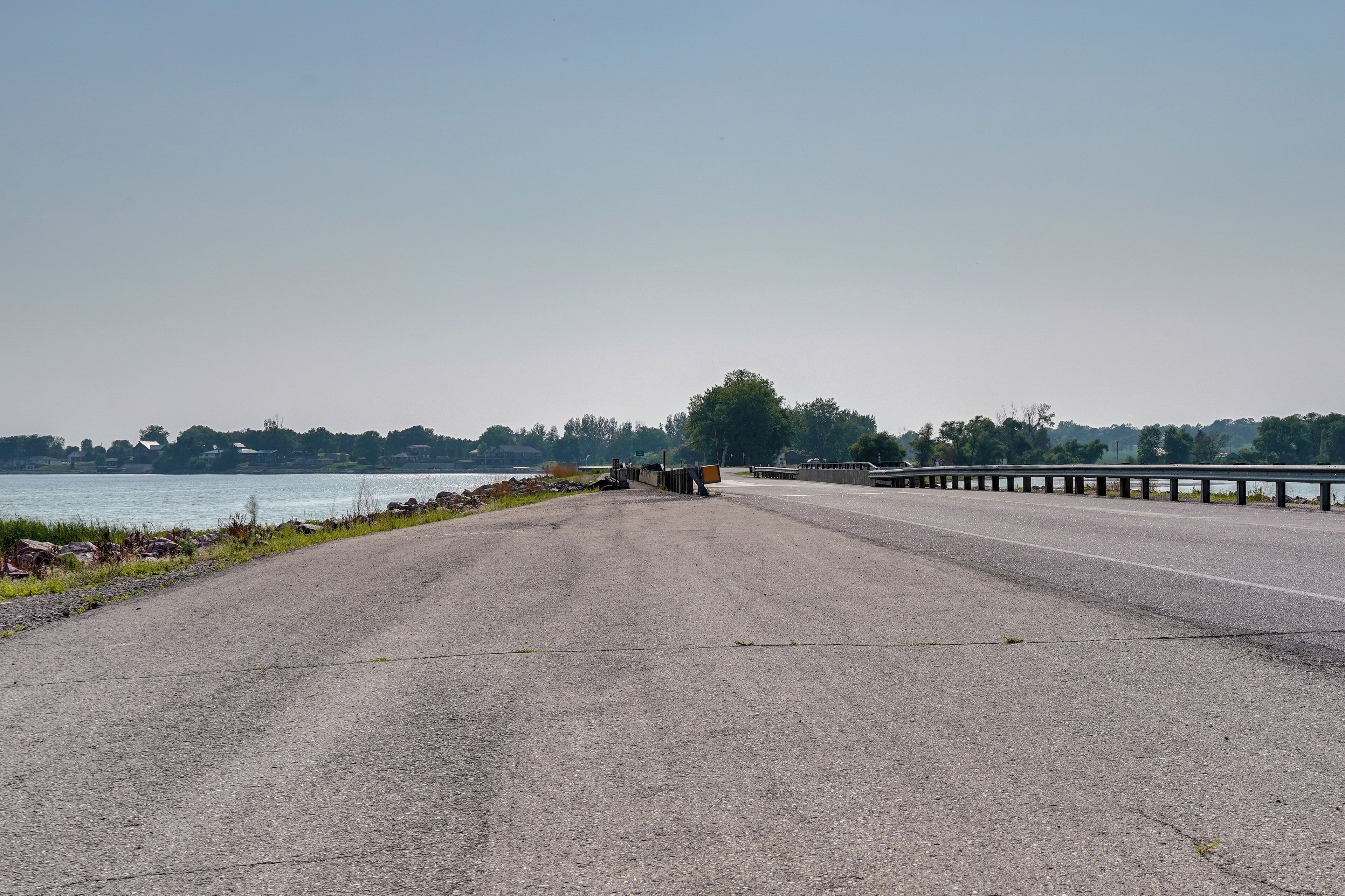 Une route asphaltée bordée d’une glissière de sécurité, un plan d’eau et une ligne d’arbres à l’horizon.