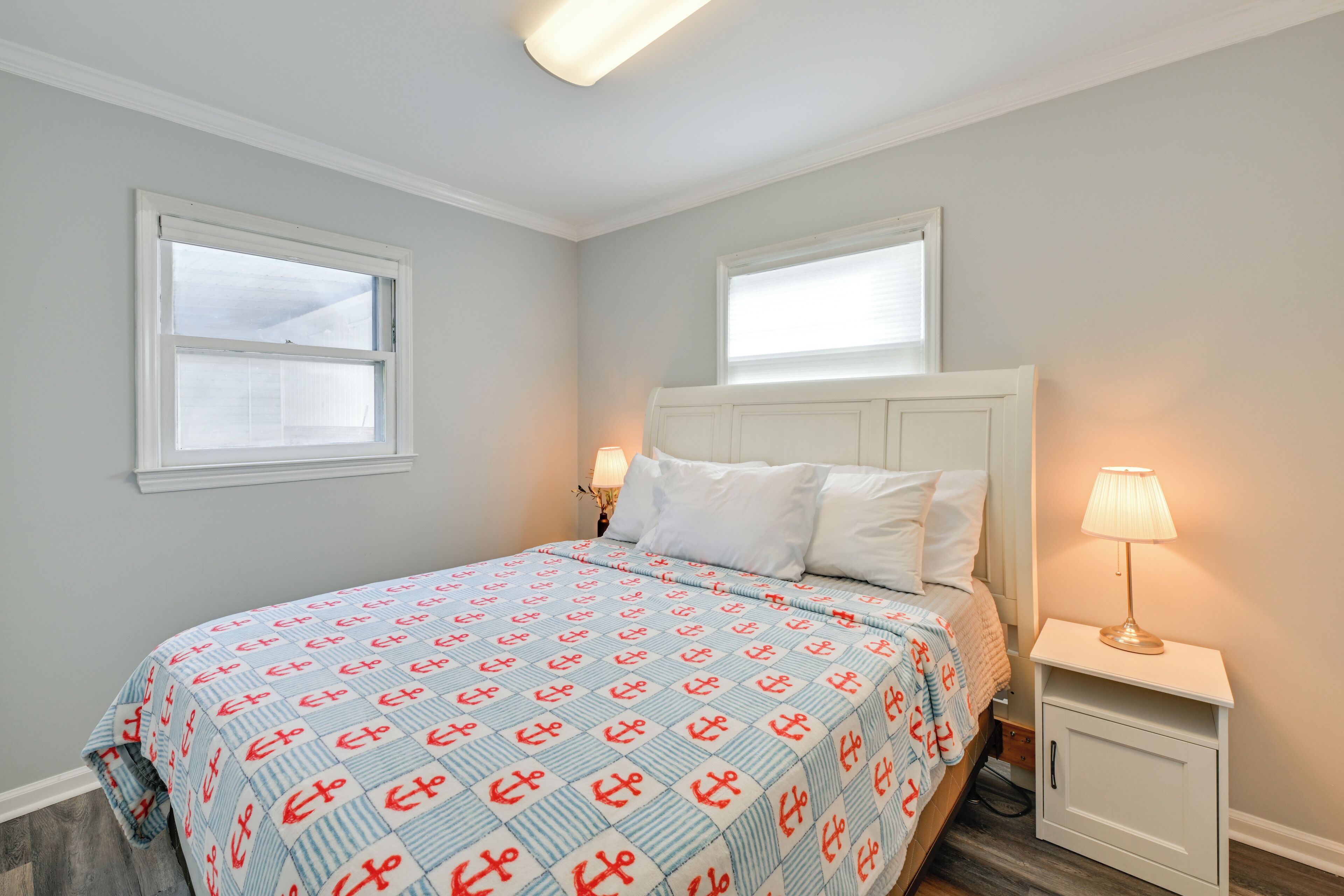 A bedroom with a bed, two windows, a nightstand with a lamp, and a small cabinet.