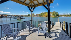 A covered deck with Adirondack chairs overlooking a lake, wooden dock, and distant buildings. - Spacious Lake Norman Retreat w/ Boat Ramp! (Sherrills Ford)