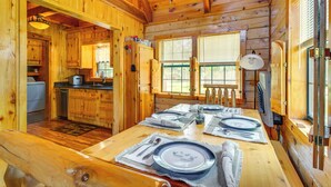 Interior de una cabaña de madera con una mesa de comedor para cuatro, una zona de cocina y una ventana con persianas.