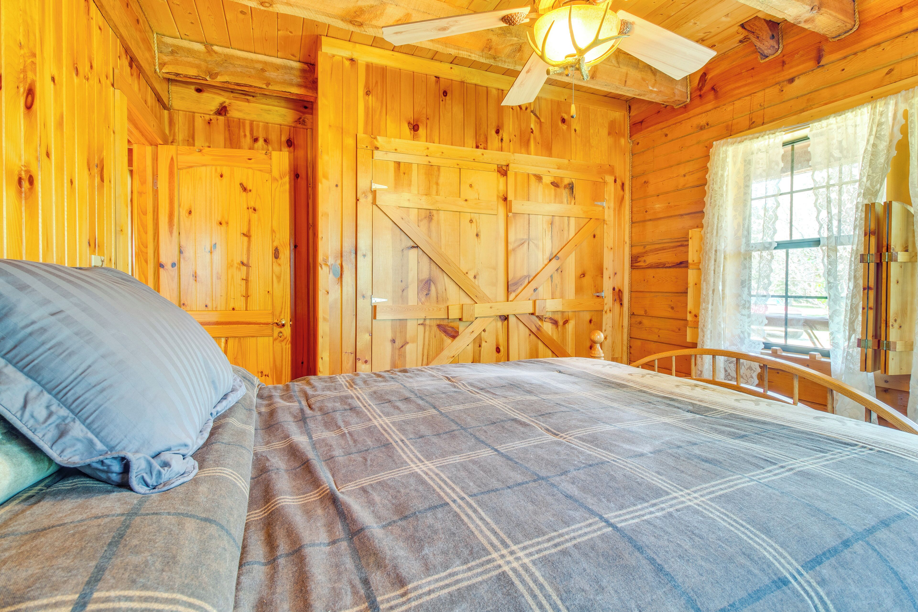 A wooden cabin room with a bed, a window with curtains, and a ceiling fan.