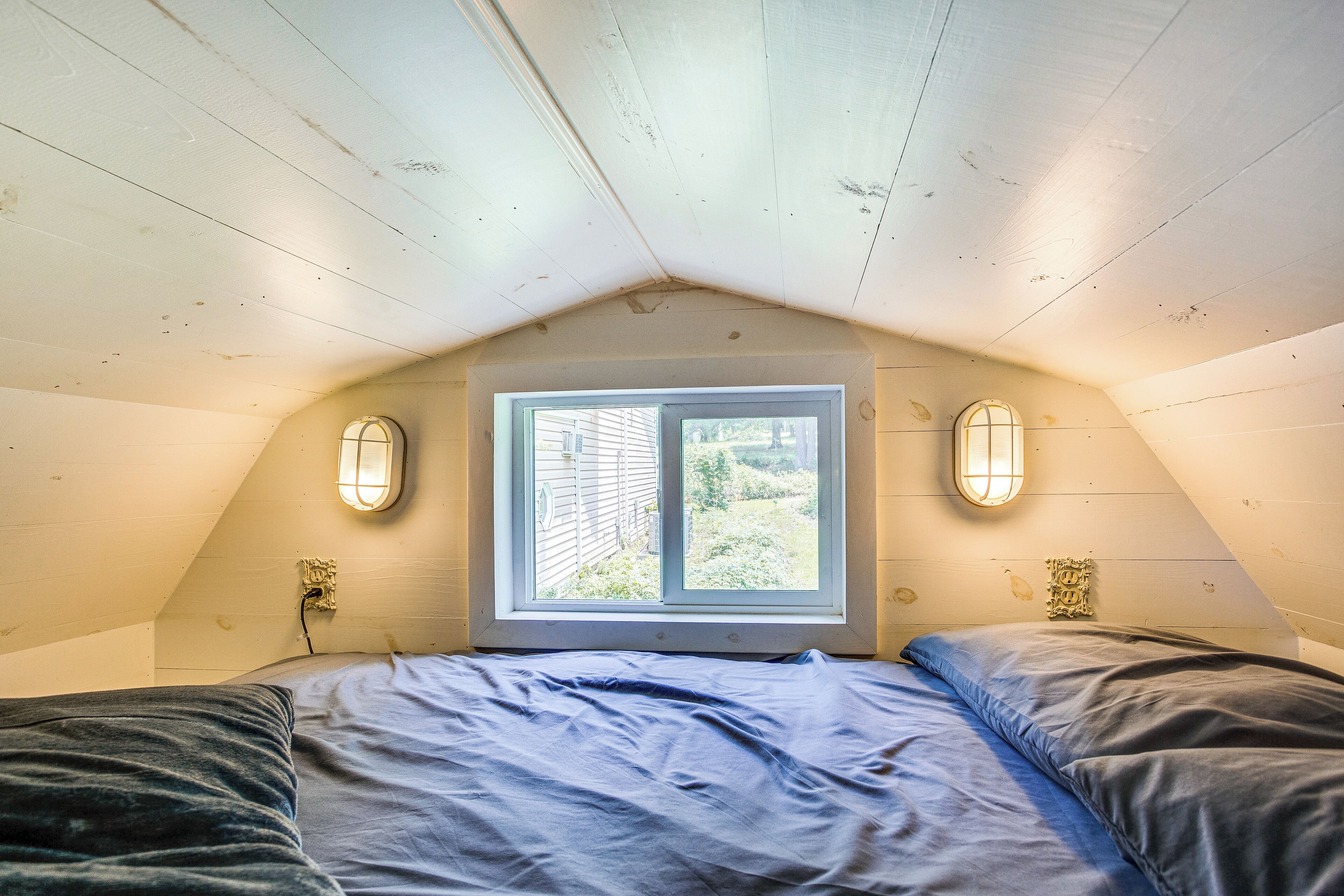 Un dormitorio con techo inclinado, una ventana con vista al exterior y dos lámparas fijadas a la pared.