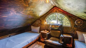 Un dormitorio ático acogedor con techo inclinado de madera, un ventanal grande, un sillón de cuero, una mesita pequeña y una cama con ropa de cama a cuadros.