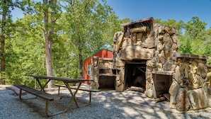 Un espacio exterior rústico con chimenea de piedra, una mesa de picnic y un edificio rojo en un entorno boscoso.