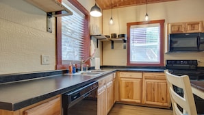 Una cocina con armarios de madera, lavavajillas, microondas y estufa. Hay dos ventanas con persianas, una lámpara colgante y una encimera con fregadero.