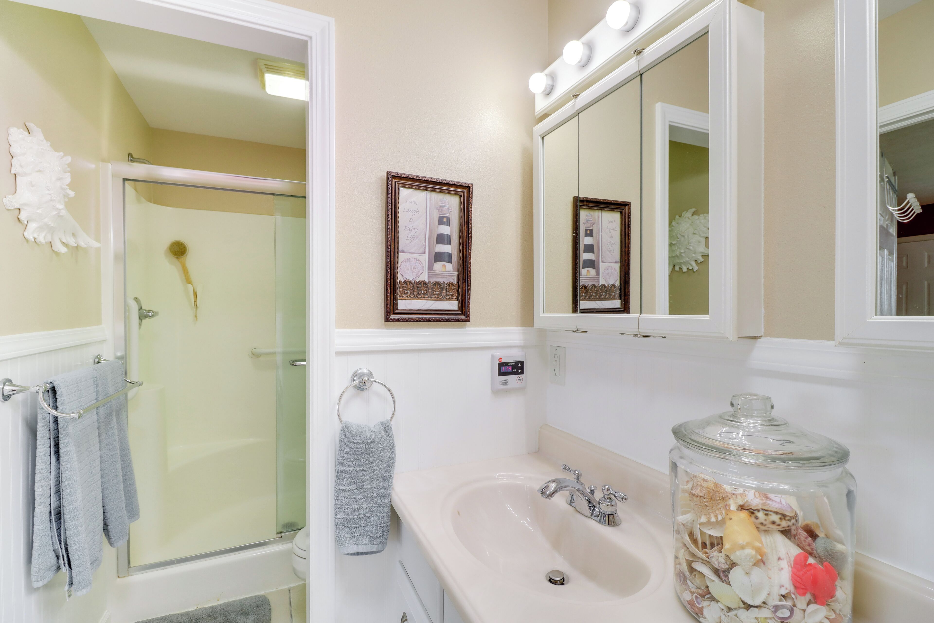 A bathroom with a glass shower enclosure, a sink with a jar of seashells, and a framed picture on the wall.