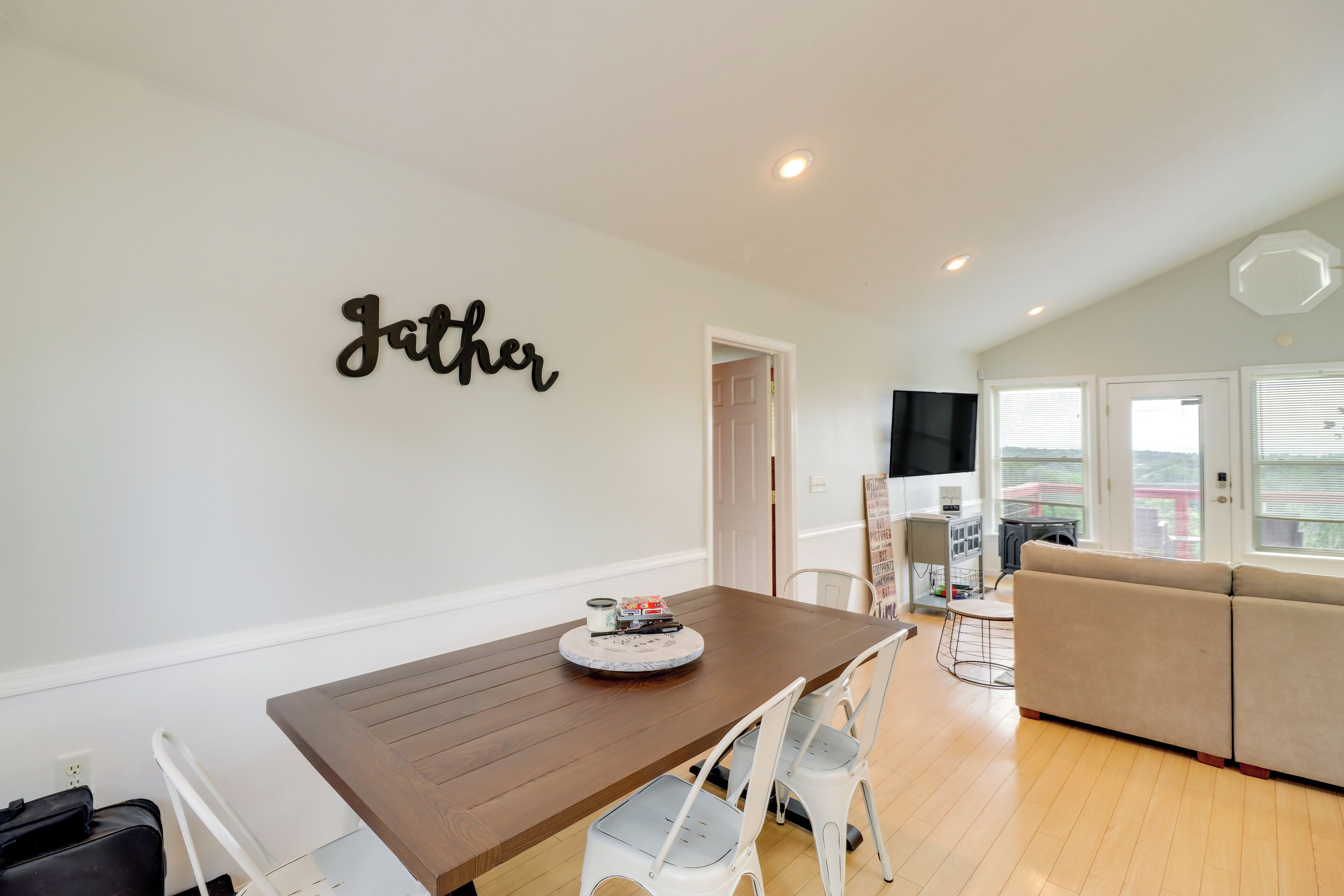 A modern living room with a brown dining table, white chairs, a beige sofa, and a wall-mounted sign that reads "Gather".