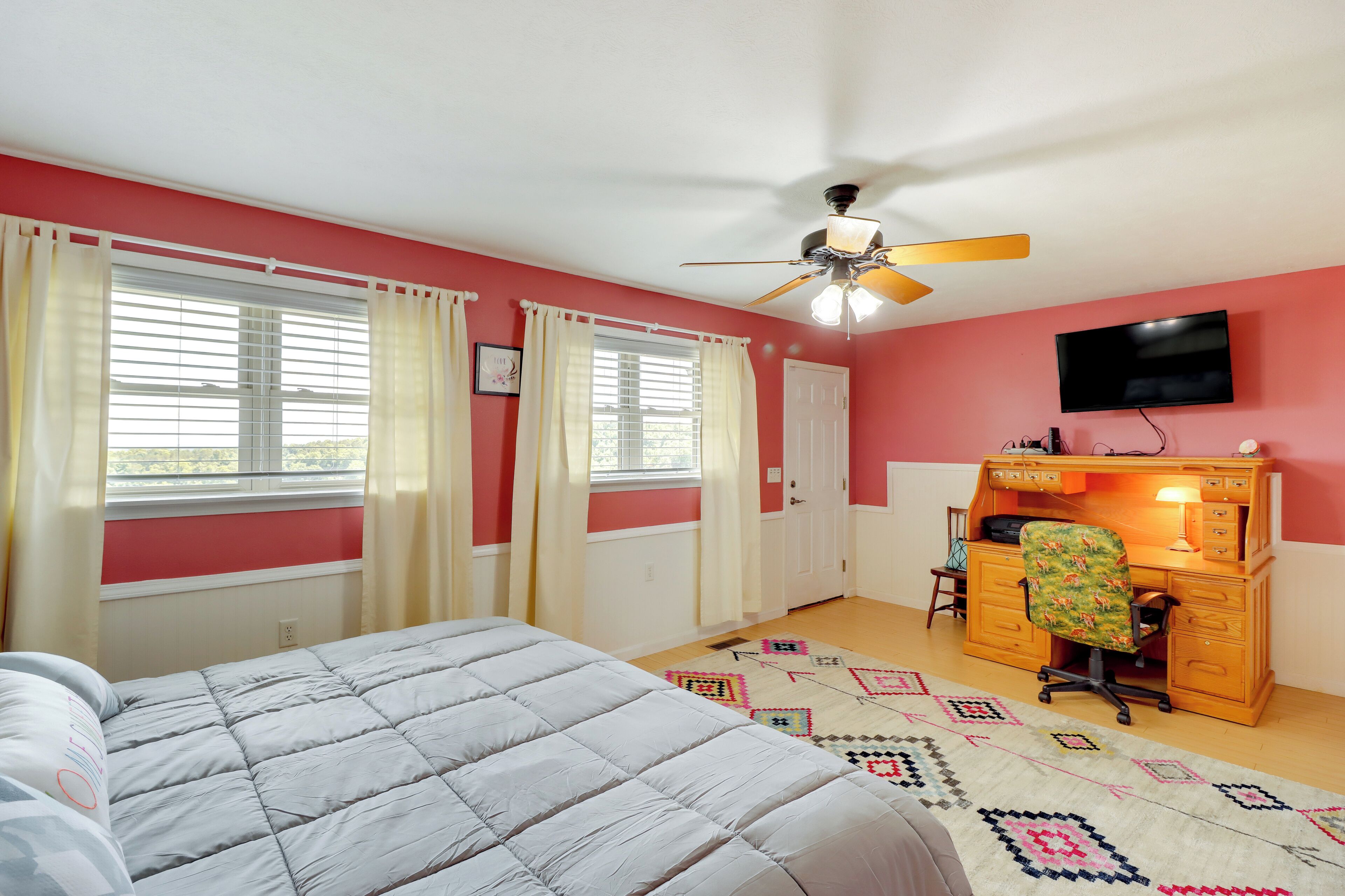 A bedroom with a bed, a desk with a chair, a ceiling fan, and a TV on the wall.