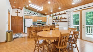 A wooden-floored dining area with a large round table and chairs, a kitchen with wooden cabinets, and a view of greenery through a door. - Serene Yellville Retreat w/ Hot Tub on 85 Acres (Yellville)