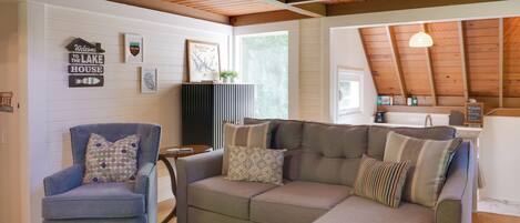 A cozy living room with a sectional sofa, a blue armchair, and a glass coffee table. There are wall decorations including a 'Welcome to the Lake House' sign and a framed picture.