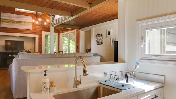 A modern kitchen with a white countertop, stainless steel sink, and a built-in oven. There is a grey sofa in the background and a large window providing natural light. - Cozy Tennessee Cabin w/ Deck, Grill & Fire Pit! (Tullahoma)