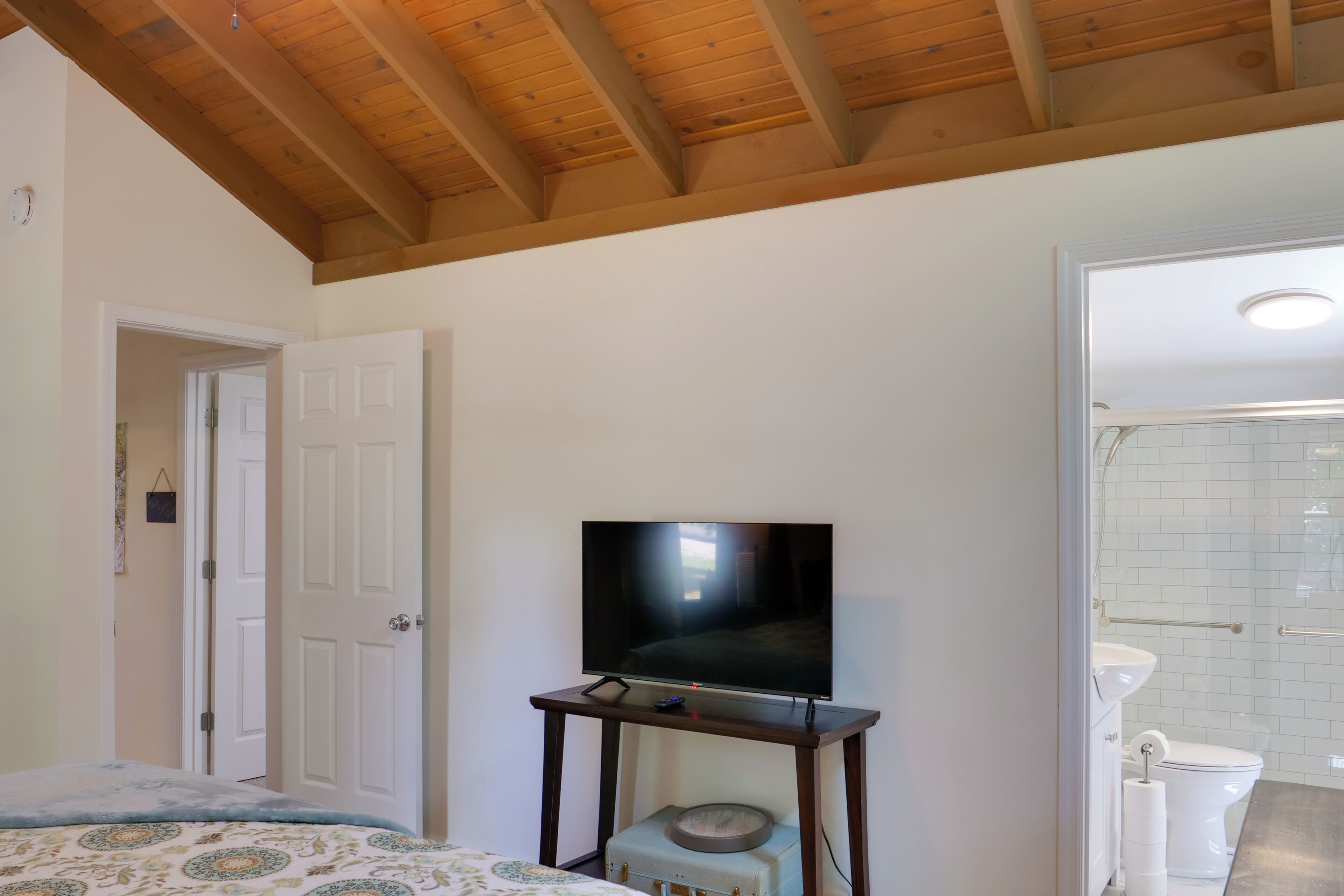 A room with a wooden ceiling, a television on a stand, a bed with a patterned bedspread, and a bathroom with a white toilet and tiled walls.
