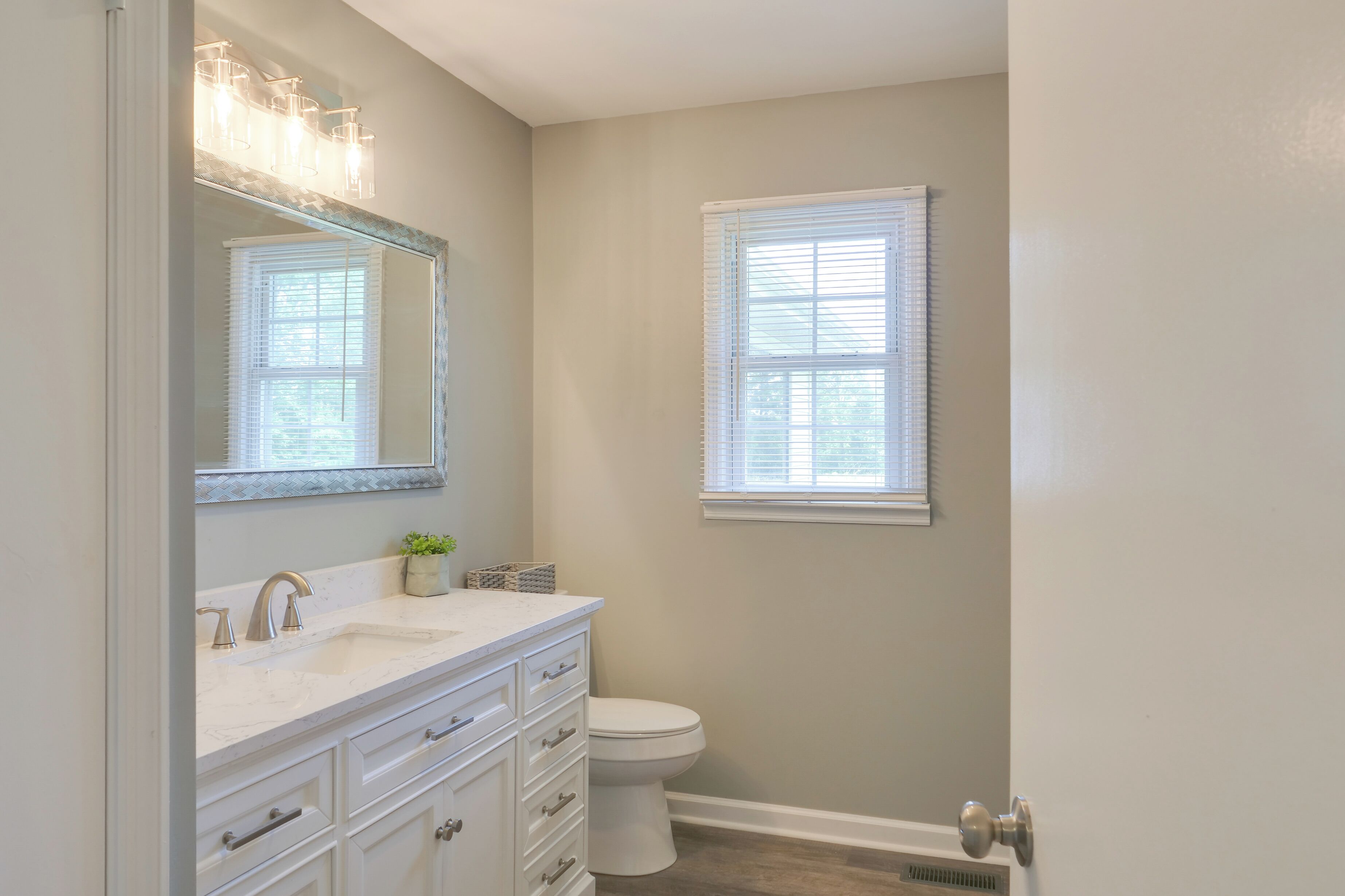 A bathroom with a white vanity, a mirror, a sink, a toilet, and a window with blinds.