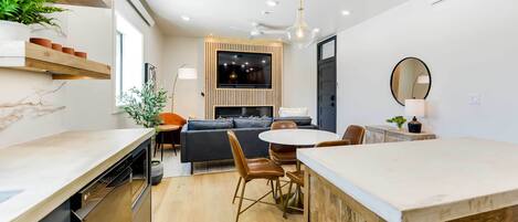 Een moderne keuken met een inbouwoven, een eethoek met een ronde tafel en stoelen, en een zithoek met een bank en een televisie.