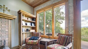 A cozy room with a brick fireplace, wooden shelves, two upholstered chairs, and a large window offering a view of trees and mountains.