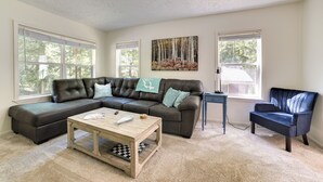 A living room with a sectional sofa, a coffee table, and a chair. There are two windows with blinds, a painting on the wall, and a lamp on a side table. - Walloon Lake Getaway w/ Fire Pit - Walk to Water! (Walloon Lake)