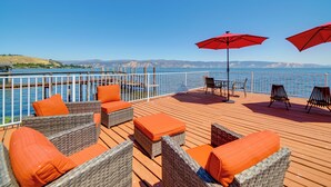 A wooden deck with wicker furniture and red cushions, overlooking a body of water with mountains in the distance.