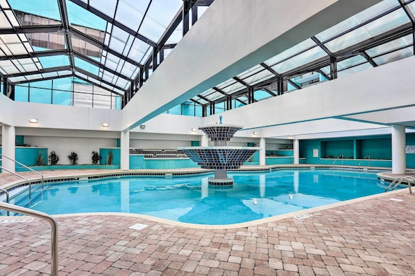 An indoor swimming pool with a central fountain, surrounded by a tiled deck and glass ceiling.