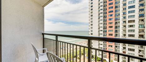 A balcony with white plastic chairs, overlooking a beach and a high-rise building with balconies.